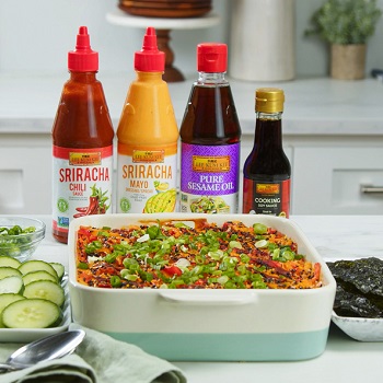 An ceramic baking dish containing a fully cooked spicy sushi bake casserole with imitation crab, surrounded by toppings and LKK sauces