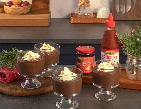Glass serving cups containing chili chocolate pudding topped with whipped cream next to bottles of LKK sauces