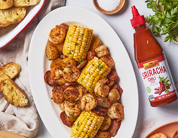 White serving platter holding a sheet pan shrimp boil, alongside garlic bread and Lee Kum Kee sriracha chili sauce