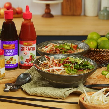 Two bowls of shortcut Beef Pho surrounded by Lee Kum Kee products, lime wedges, and other traditional toppings
