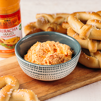 A bowl of spicy pub cheese dip from Lee Kum Kee’s recipe on a board with a stack of pretzels and beside a bottle of Lee Kum Kee Sriracha Mayo