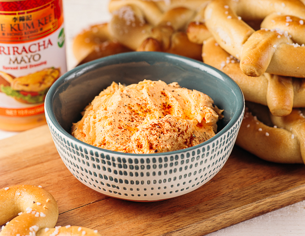 A bowl of spicy pub cheese dip from Lee Kum Kee’s recipe on a board with a stack of pretzels and beside a bottle of Lee Kum Kee Sriracha Mayo
