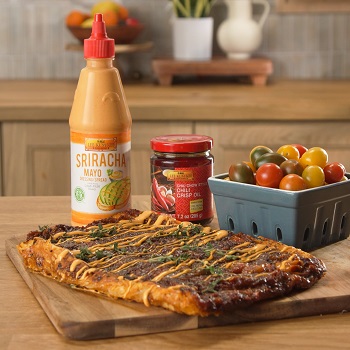 Tomato Tarte Tatin recipe on a wooden board, accompanied by a basket of tomatoes, Lee Kum Kee Sriracha Mayo, and Lee Kum Kee Chiu Chow Chili Crisp Oil for added flavor.